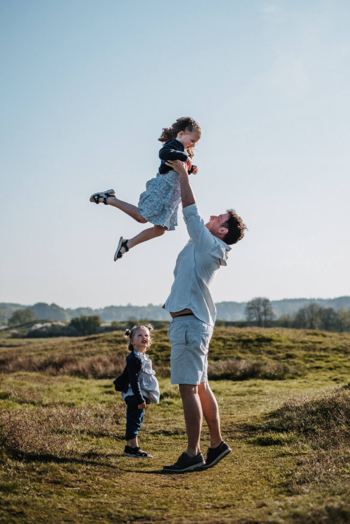 Vater mit zwei Kindern vor blauem Himmel. Er hebt seine Tochter hoch, beide lachen