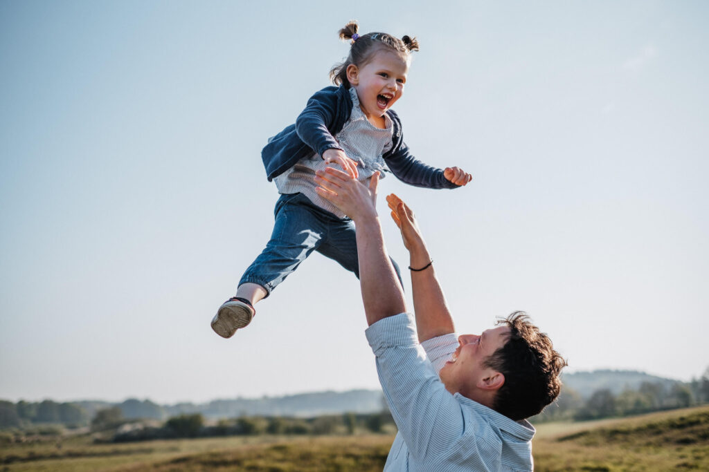 Vater wirft lachende Tochter in die Luft vor blauem Himmel, Familienfotografie in Flensburg.