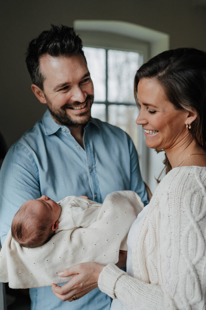 Stolze Eltern mit Baby im Arm lachen bei einem Neugeborenen Shooting in Flensburg.