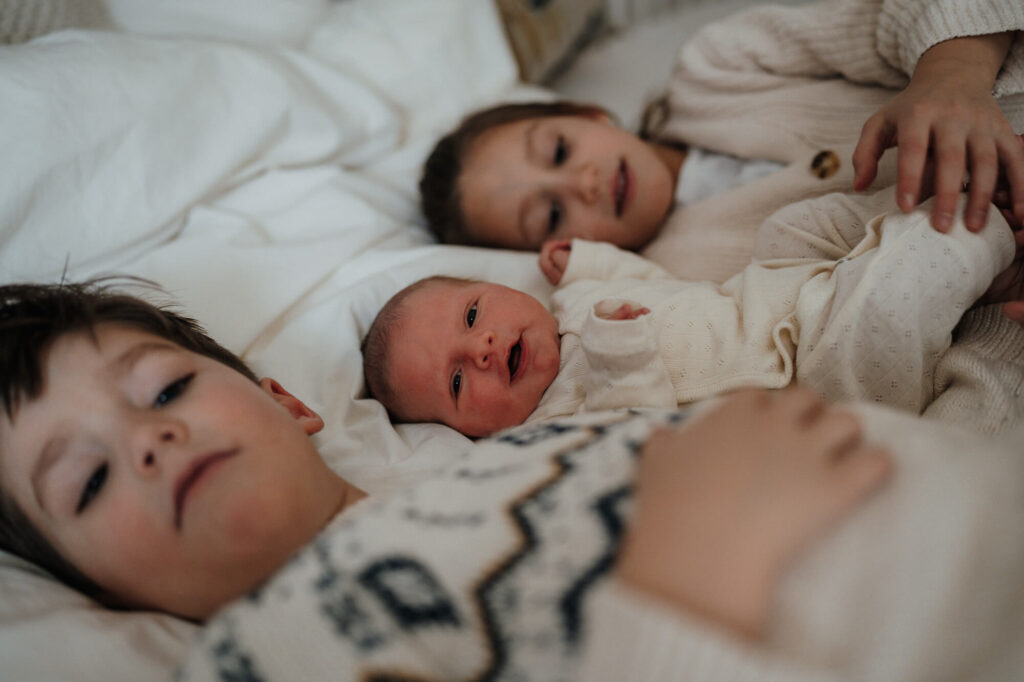 Zwei ältere Geschwister mit ihrem Neugeborenen im Bett – Neugeborenen Fotografin Flensburg