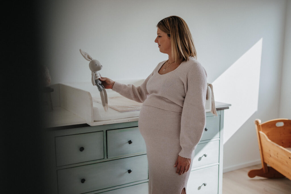 Frau mit Babybauch hält einen Stoffhasen hoch und steht vor einer Wickelkommode