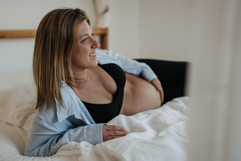 schwangere Frau liegt auf dem Bett und hält lachend ihren Babybauch