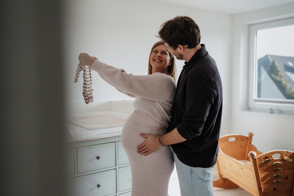 Pärchen bei einem Babybauch Shooting in Flensburg lacht sich an und hält einen kleinen Body hoch